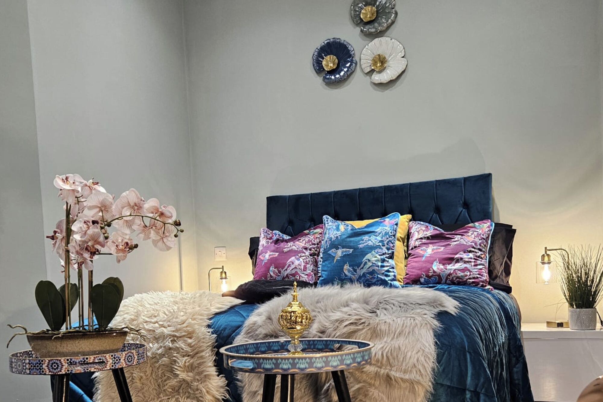 A bedroom with a blue upholstered headboard, a fluffy white rug, a decorative bowl, and a potted plant.