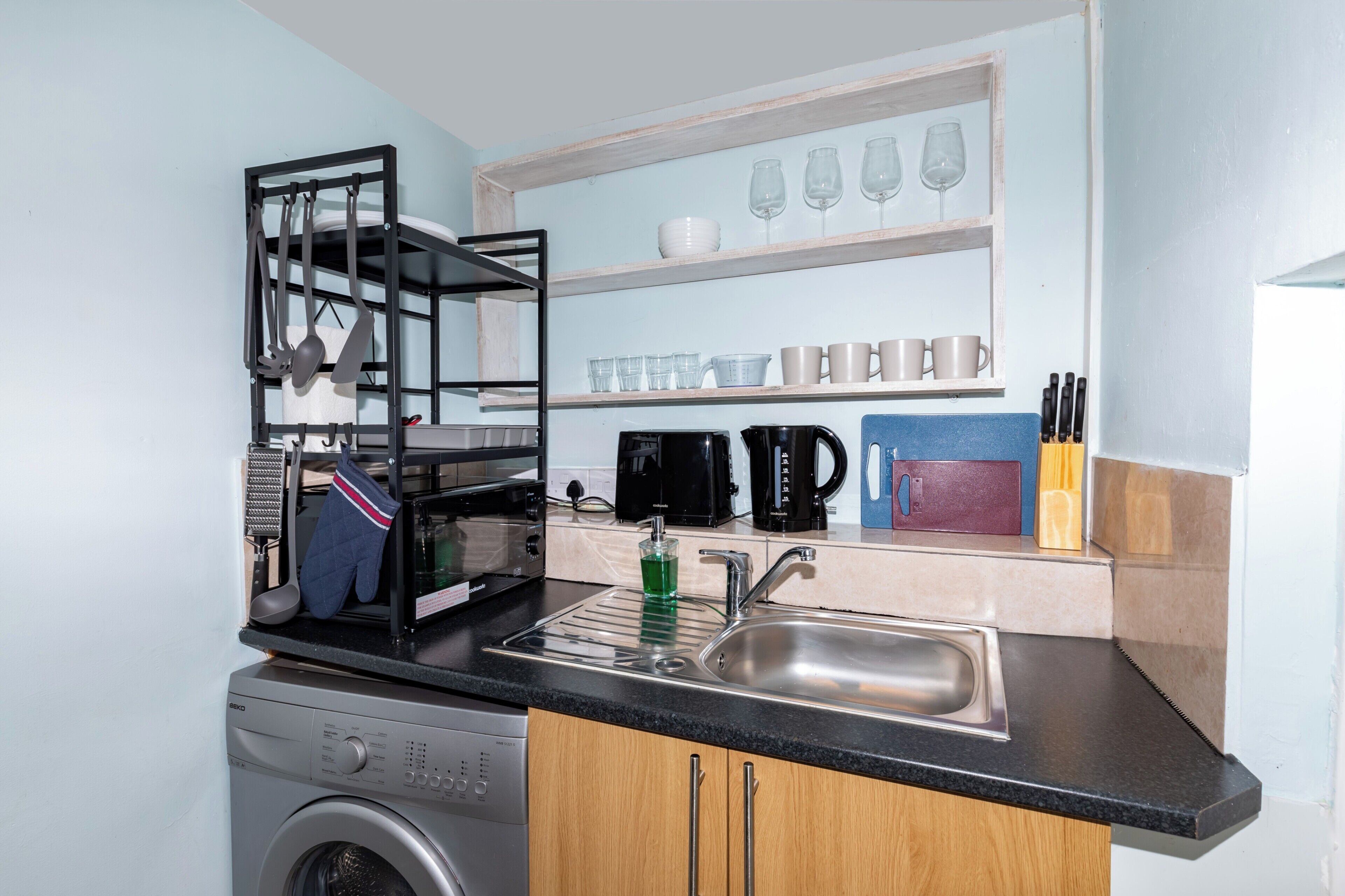 A compact kitchen with a washing machine, a sink, a drying rack, and shelves with glassware and kitchen utensils.