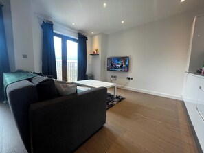 A modern living room with a sofa, a TV mounted on the wall, and a balcony with blue curtains. - Grosvenor Rd Studio - Sleeps 4, Pet Friendly, Wifi (St Albans)