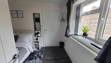 Una habitación de hotel estrecha con una cama, un banco, un espejo, un radiador y una ventana con una planta.