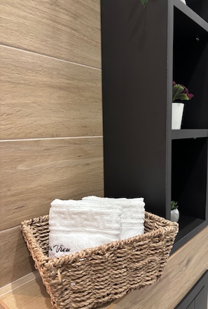Ein Korbwicker-Korb mit gefalteten Handtüchern auf Holzboden neben einer dunklen Wand und einem Regal mit einer kleinen Pflanze.