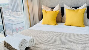A hotel room with a large bed, two yellow pillows, a white towel, and a view of a cityscape through a window. - The Bullring Studio in Birmingham City Centre (Birmingham)