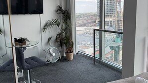 A modern room with a large window, a flat-screen TV, a small table with a basket, two potted plants, a grey chair, and a standing fan. - The Bullring Studio in Birmingham City Centre (Birmingham)