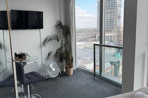A modern room with a large window, a flat-screen TV, a small table with a basket, two potted plants, a grey chair, and a standing fan. - The Bullring Studio in Birmingham City Centre (Birmingham)