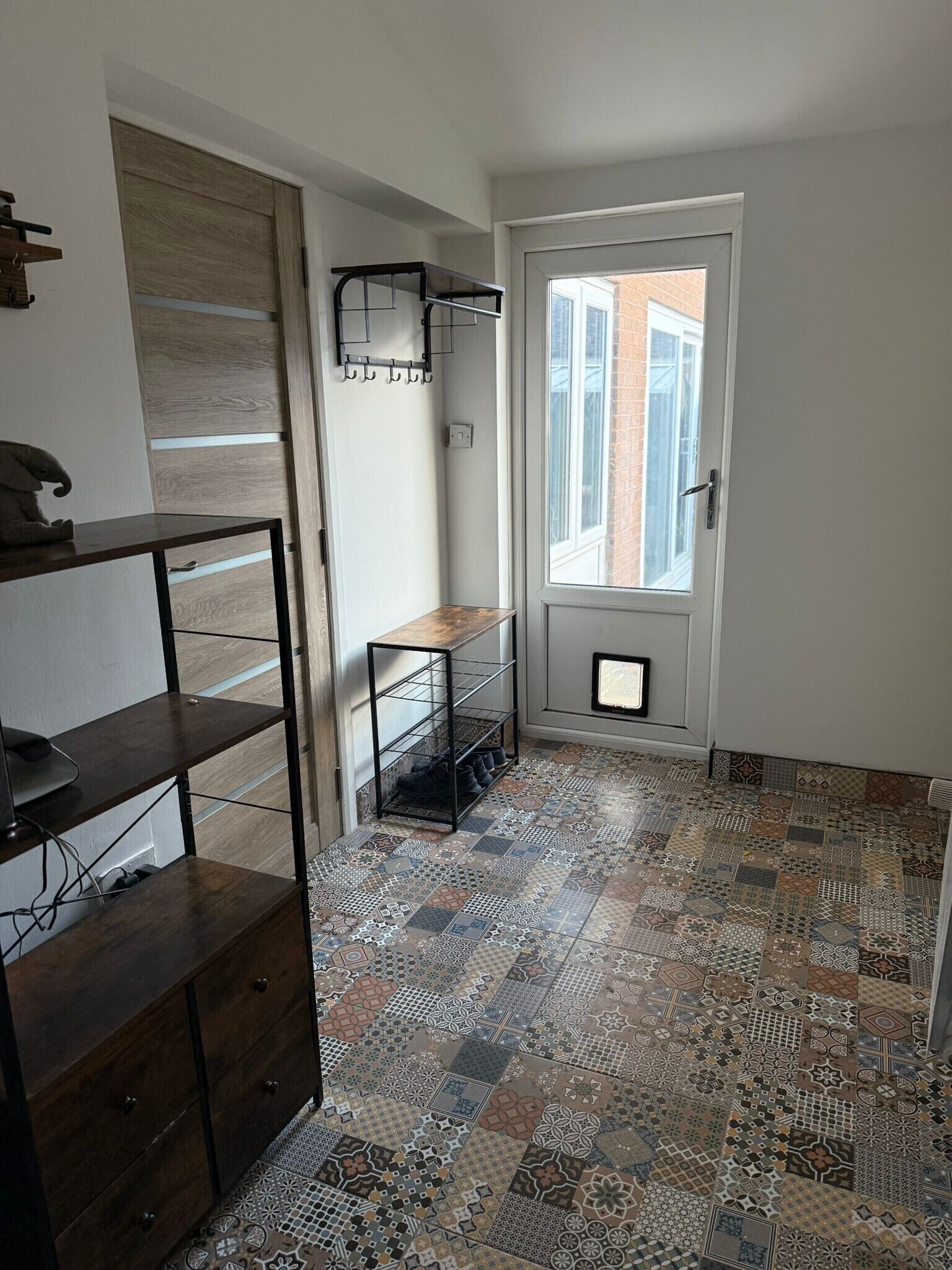 A room with a patterned floor, a wooden cabinet, a shoe rack, and a window with a view of a brick building.