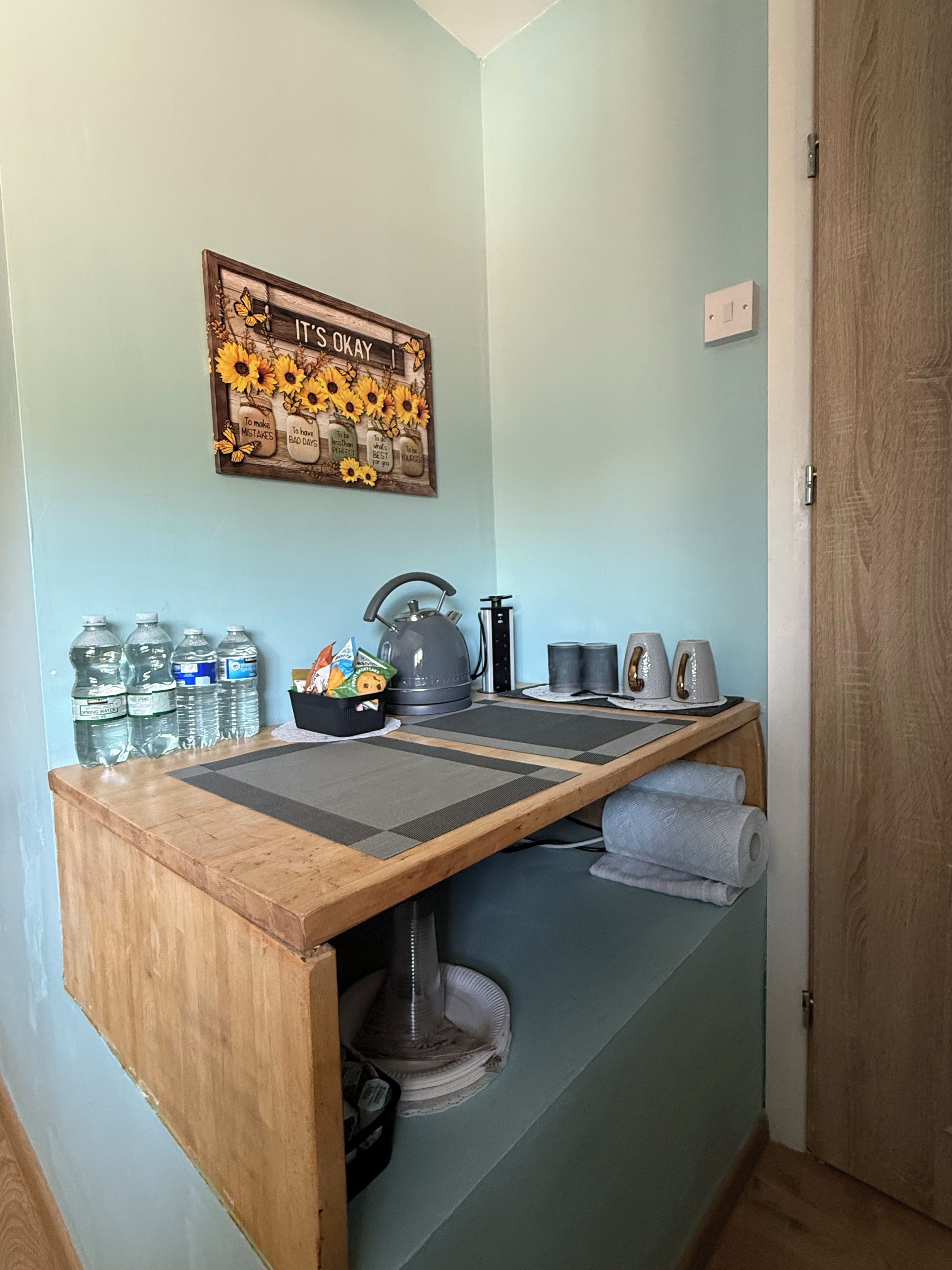 A compact kitchen with a wooden countertop, a kettle, a kettle pot, and a coffee maker. There are water bottles and a small bowl on the counter.