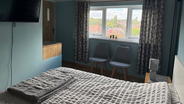 Un dormitorio con una cama, un televisor instalado en la pared, dos sillas, una ventana con cortinas y una mesita de noche con lámpara.