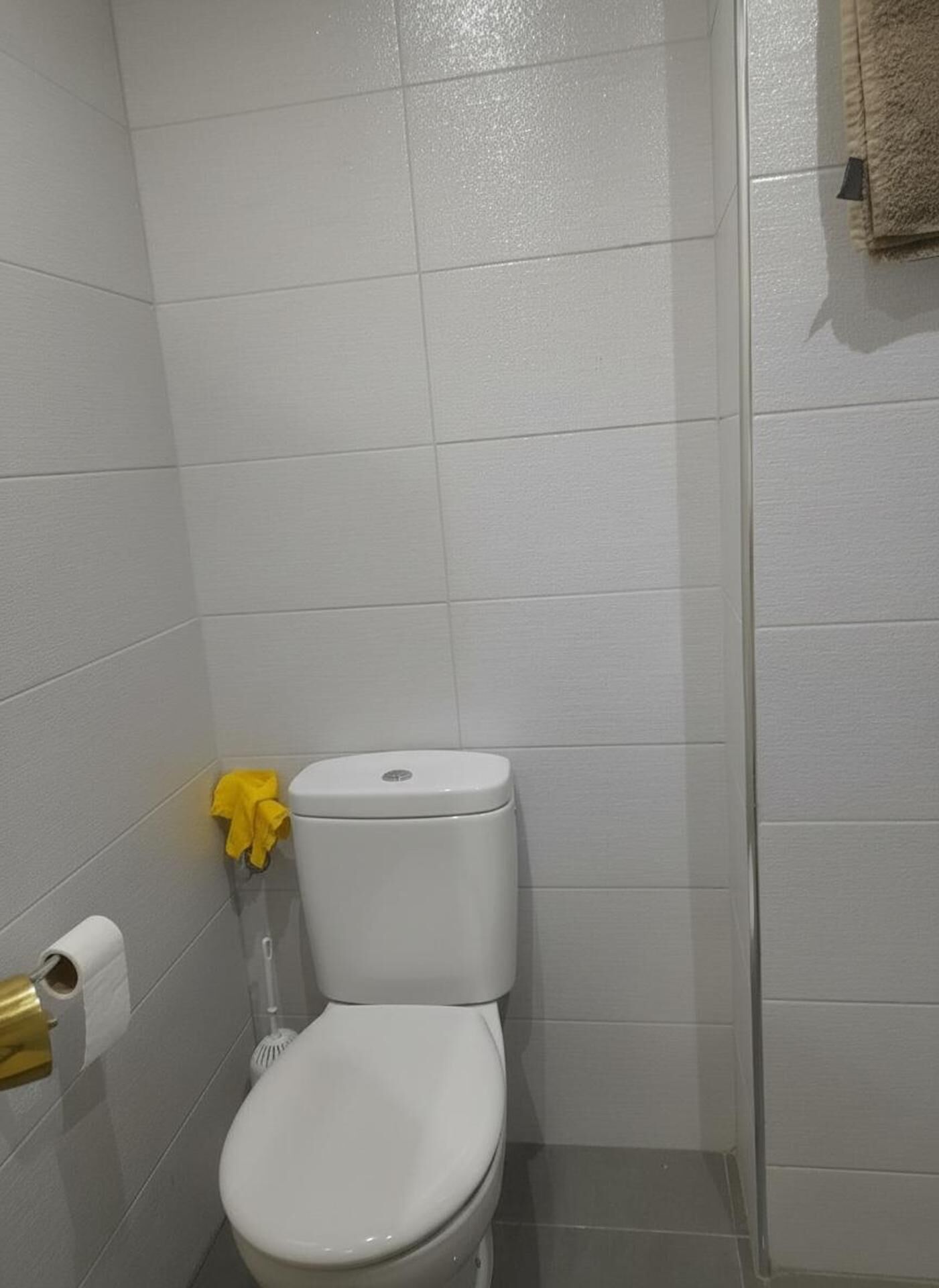 Ein kleines Badezimmer mit weißen Fliesen, einer Toilette, einem gelben Handtuch und einem Toilettenpapierhalter.