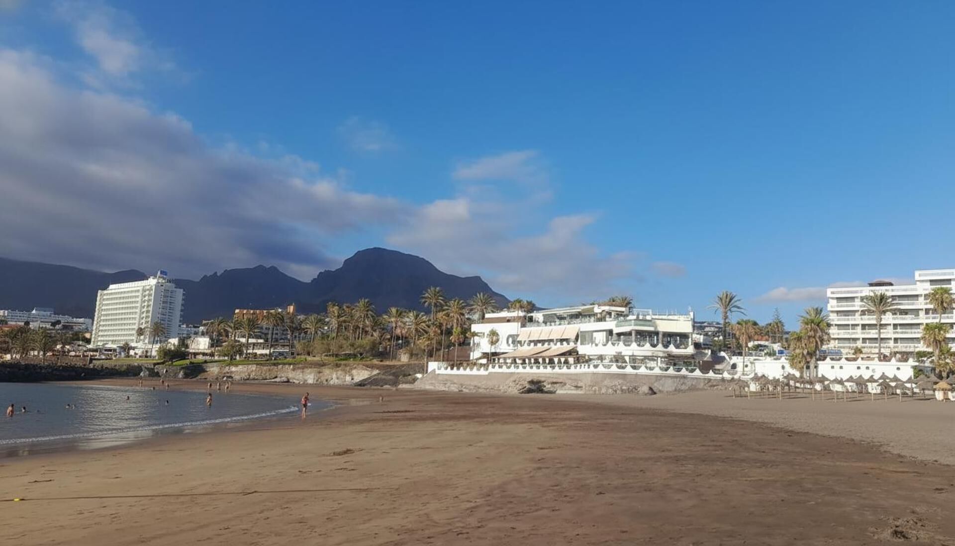 A beach with buildings, palm trees, and mountains in the background.