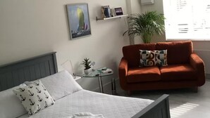 En pænt redt seng med puder, et lille natbord med en plante, en rød sofa med pude med mønster, og en vægmonteret hylde med bøger og et ur.