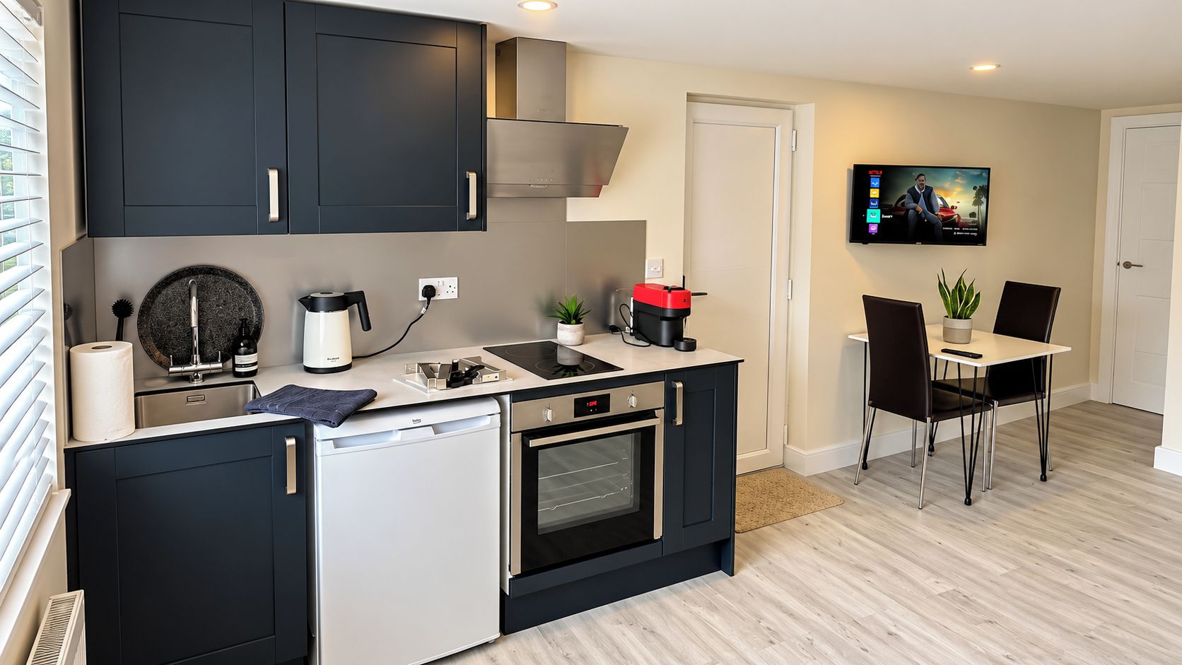 A compact kitchen with a dishwasher, oven, and coffee maker, a dining area with chairs, and a TV mounted on the wall.