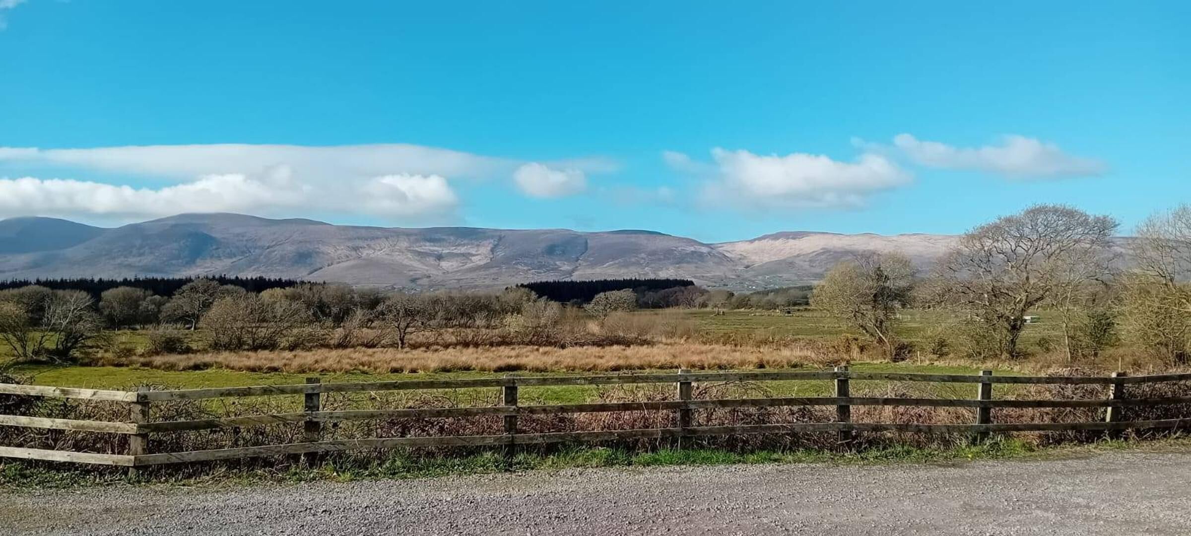 Paisatge rural amb una tanca de fusta, camps de gespa i muntanyes llunyanes sota un cel serè.