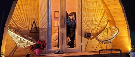 Estructura de madera acogedora, semicerrada, con puerta, dos sillas y una mesa en el exterior.