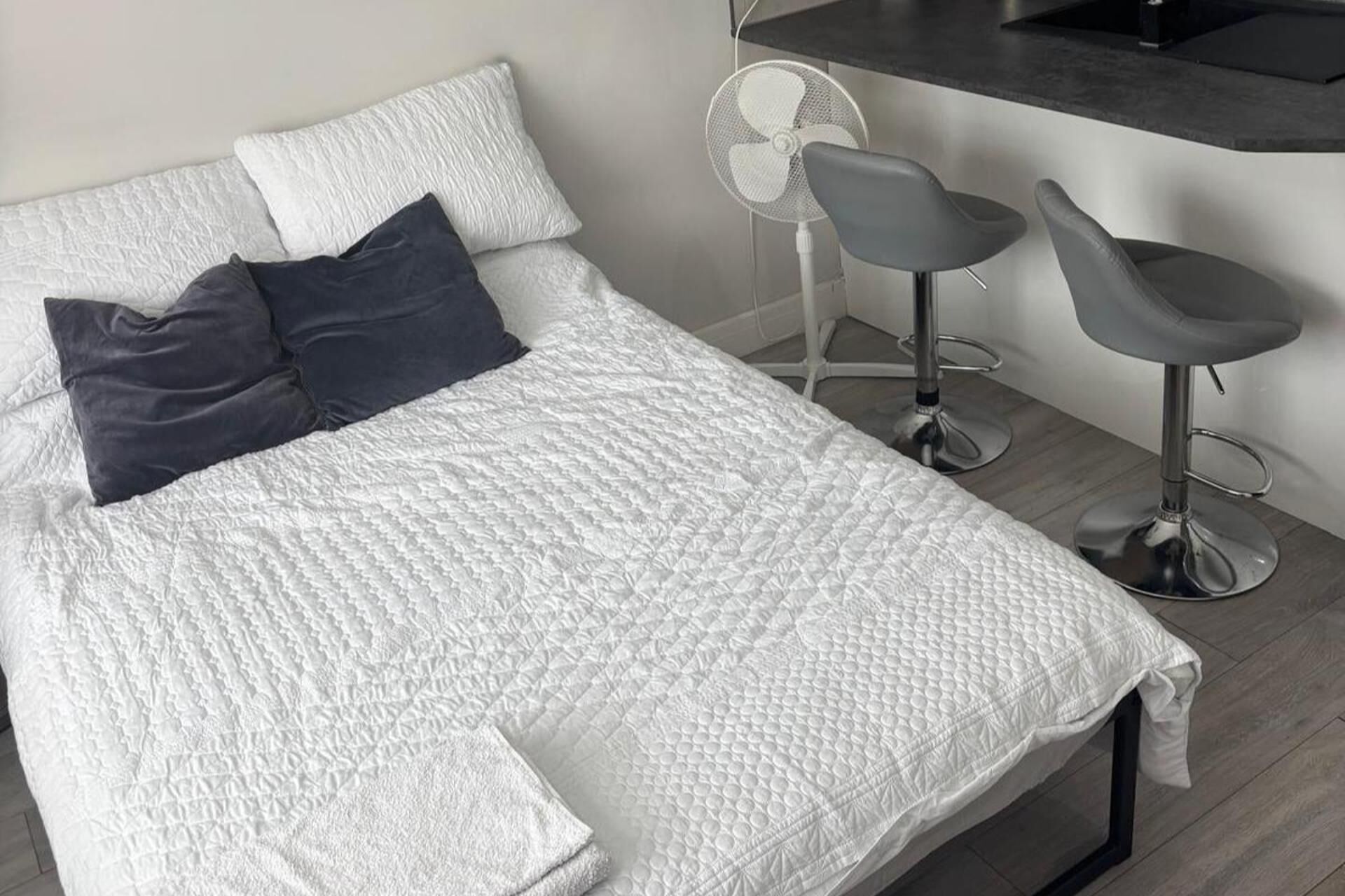 Un lit avec une couette blanche, deux oreillers, un petit ventilateur et deux tabourets de bar gris.