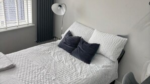 Een keurig opgemaakt bed met een witte quilt, twee donkere kussens en een bedlampje.