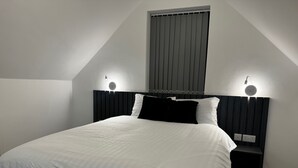 A bedroom with a bed, two bedside lamps, a window with blinds, and a wall-mounted power outlet. - Studio Flat In London (Bexleyheath)