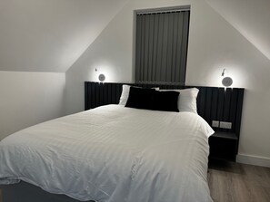 A bedroom with a bed, two bedside lamps, a window with blinds, and a wall-mounted power outlet. - Studio Flat In London (Bexleyheath)