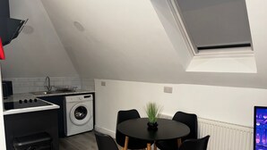 A compact kitchen with a washing machine, a dining table with chairs, and a small trash bin. - Studio Flat In London (Bexleyheath)