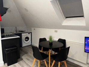 A compact kitchen with a washing machine, a dining table with chairs, and a small trash bin. - Studio Flat In London (Bexleyheath)