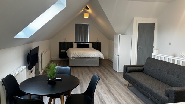 A modern loft apartment with a skylight, a bed, a refrigerator, a gray sofa, and a dining area with a table and chairs. - Studio Flat In London (Bexleyheath)