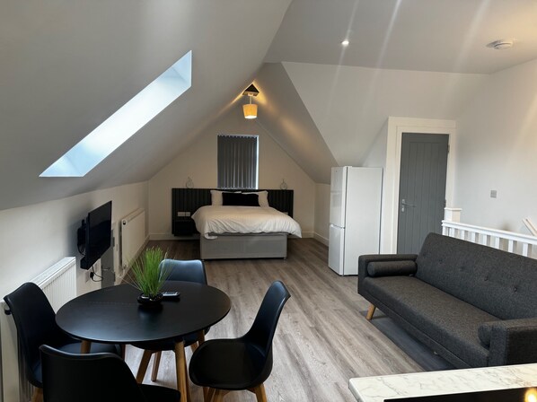 A modern loft apartment with a skylight, a bed, a refrigerator, a gray sofa, and a dining area with a table and chairs. - Studio Flat In London (Bexleyheath)