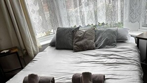 A neatly made bed with pillows and rolled towels, a window with lace curtains, and a bedside table. - Studio Sleeps 1 - Pets Welcome -free Parking (Blackpool)