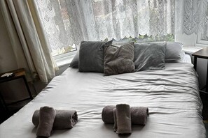 Ein ordentlich gemachtes Bett mit Kissen und gerollten Handtüchern, ein Fenster mit Spitzenvorhängen und ein Nachttisch.