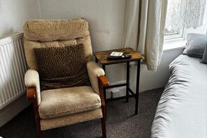 Ein Sessel mit gemustertem Kissen, ein kleiner Holztisch mit Fernbedienung und ein Bett mit Kopfkissen.