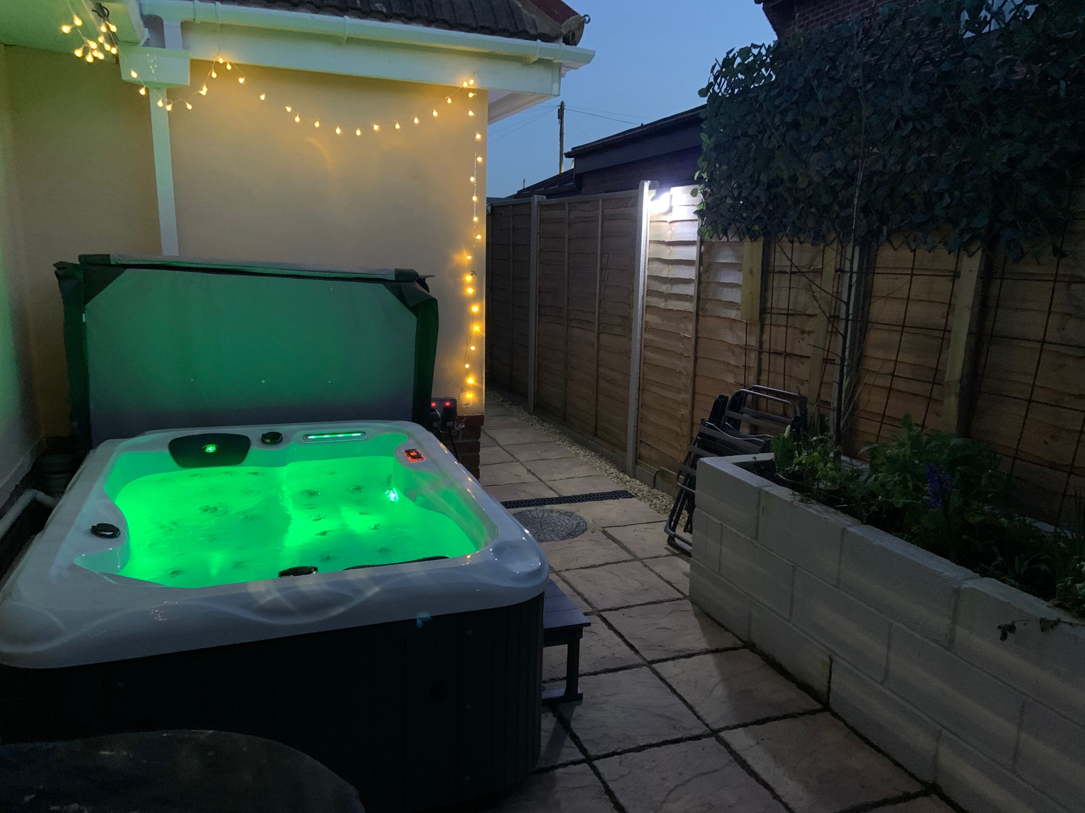 Un jacuzzi con luz verde, rodeado de un patio con azulejos y una valla de madera.