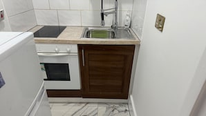 Pieni, käytännöllinen keittiö, jossa on valkoinen jääkaappi, liesi, pesuallas ja puukehikoiset kaapit.