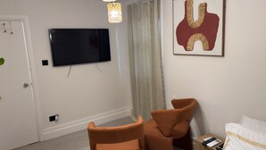 Moderni hotellihuone, jossa on seinälle asennettu taulutelevisio, suuri abstrakti maalaus, kaksi ruskeaa nojatuolia, valkoisilla lakanoilla varustettu sänky ja yöpöytä, jossa on kaukosäädin.