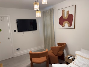 Ein modernes Hotelzimmer mit einem an der Wand befestigten Flachbildfernseher, einem großen abstrakten Gemälde, zwei braunen Sesseln, einem Bett mit weißer Bettwäsche und einem Nachttisch mit Fernbedienung.