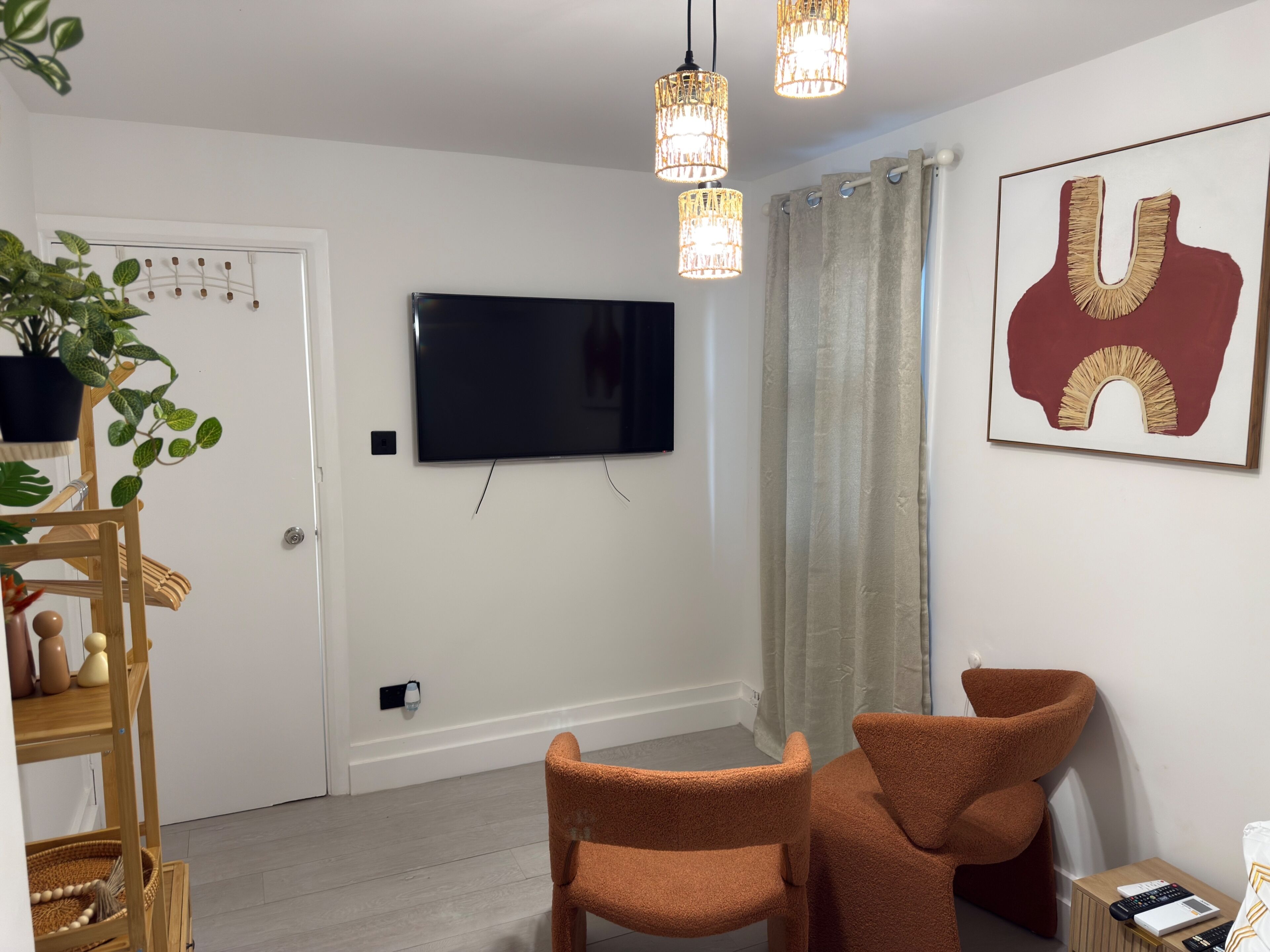 Un salon moderne avec un téléviseur à écran plat, deux fauteuils orange, une étagère en bois ornée d’objets décoratifs, une plante en pot et une œuvre d’art encadrée accrochée au mur.