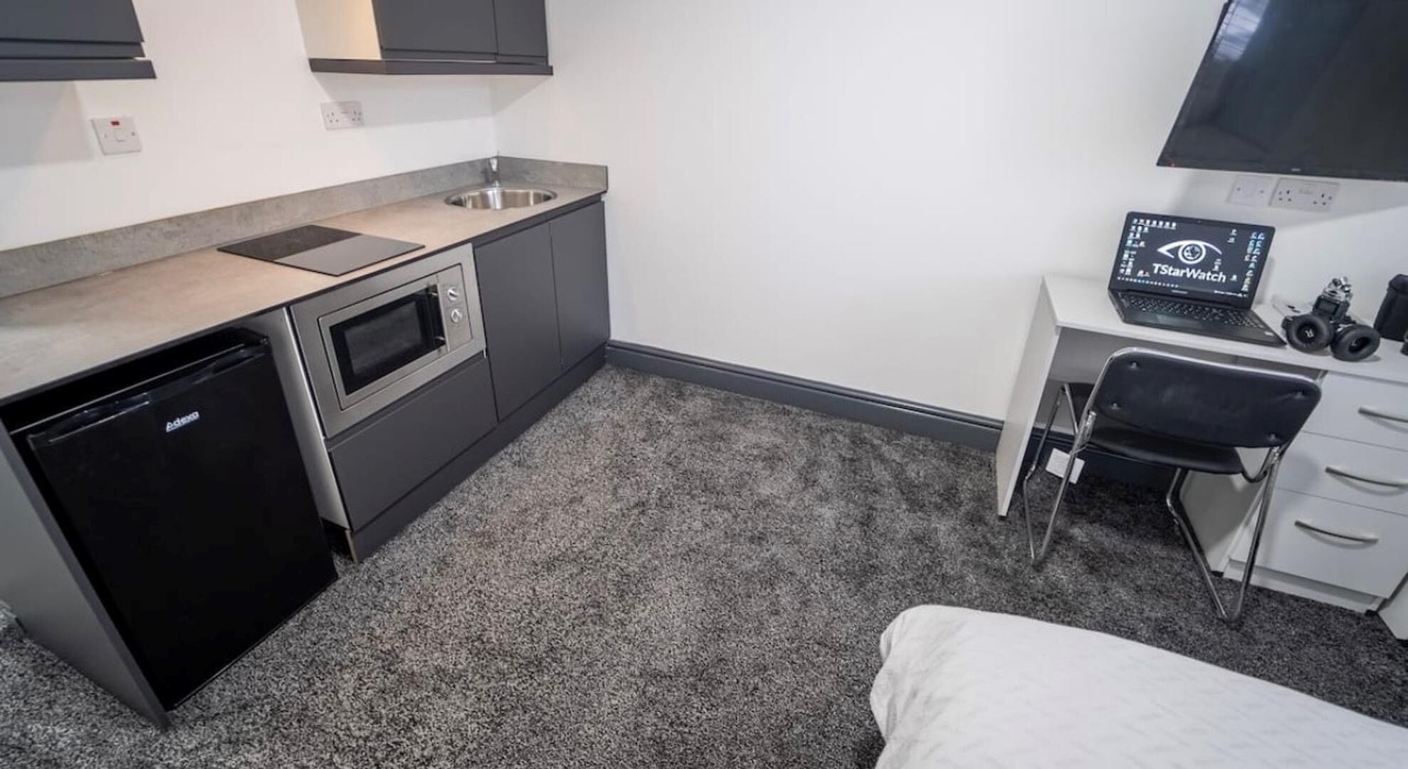 A compact kitchen with a dishwasher, oven, and a small sink. There's a desk with a computer and headphones, and a bed in the corner.