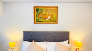 Una cama impecable con sábanas blancas y dos almohadas color beige, flanqueada por mesitas de noche con lámparas y cuadros enmarcados.