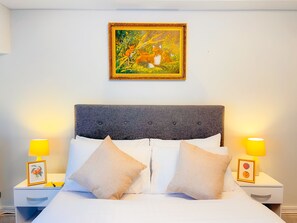 A neatly made bed with white linens and two beige pillows, flanked by nightstands with lamps and framed pictures. - London - Apartment - Riverside - Sleeps 3 - Garden (London)
