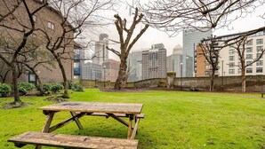 Un espacio cubierto de césped con una mesa de picnic de madera y bancos, rodeado de árboles y edificios al fondo.
