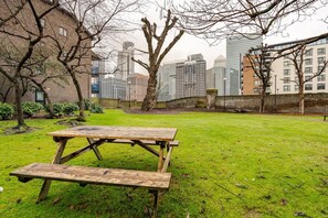 A grassy area with a wooden picnic table and benches, surrounded by trees and buildings in the background. - London - Apartment - Riverside - Sleeps 3 - Garden (London)