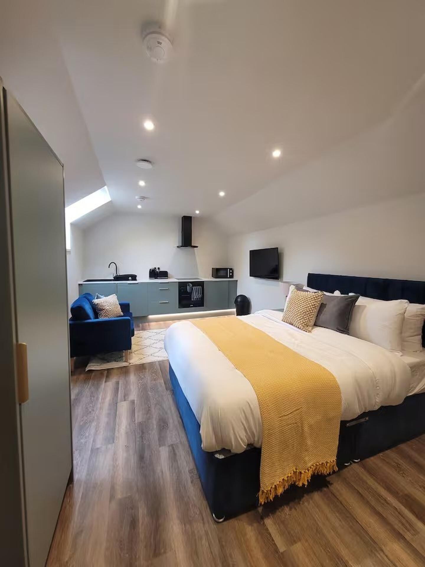 A modern bedroom with a large bed, a small kitchenette, and a TV mounted on the wall.