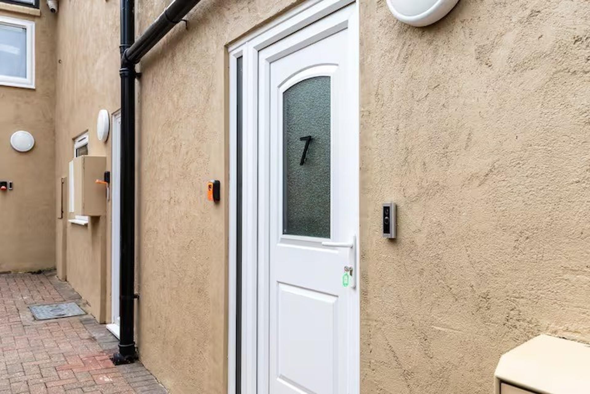 A narrow alleyway with a white door marked with the number 7, a small window, and a wall-mounted intercom.