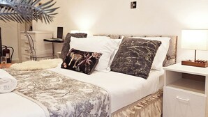 A neatly made bed with a patterned bedspread, two decorative pillows, a bedside table with a lamp, and a small drawer unit. - Studio Sleeps 3 - Free Wifi - Flat-screen Tv (London)