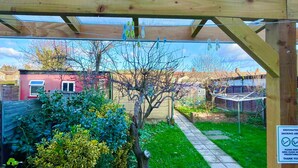 A wooden pergola structure with a garden area beneath, a pathway, and a red building in the background. - Bright Cosy Studio & Free Parking-greater London (Enfield)