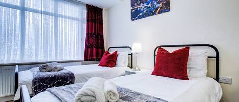 A hotel room with two beds, a window with sheer curtains, a cityscape painting, and a radiator.