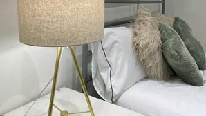 Uma cama com roupa de cama branca e almofada verde e bege, uma luminária dourada com cúpula bege e uma mesa de cabeceira branca.