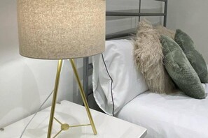 Un llit amb roba de llit blanca i un almoïna verd i beix, una làmpada daurada amb pantalla beix i una tauleta de nit blanca.