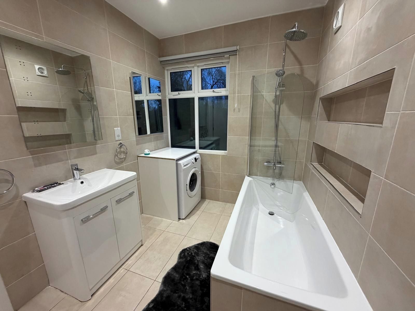 Un baño moderno con bañera, lavamanos, lavadora y ducha con mampara de cristal.