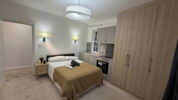 Un dormitorio con cama, mesitas de noche, armario, zona de cocina con lavabo y estufa, y una lámpara colgante.