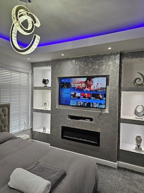 A modern bedroom with a television mounted on the wall, a bed with grey bedding, and decorative items on shelves.
