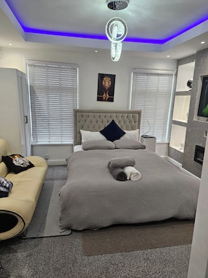 Una camera da letto moderna con una testiera trapuntata, un letto grande con biancheria grigia, un divano con cuscini a tema leonino e una TV a parete.
