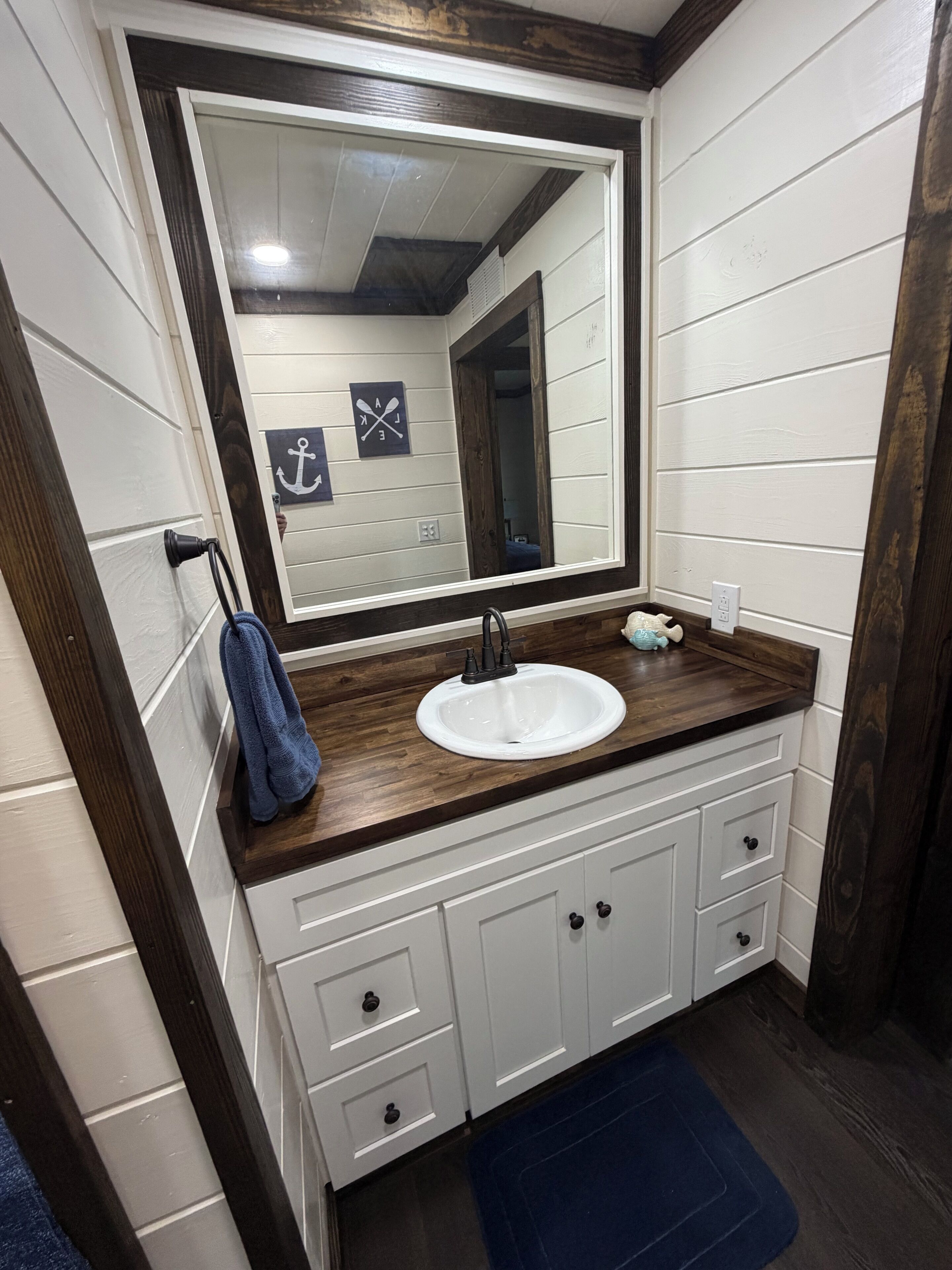 A bathroom with a white sink, dark wooden countertop, and a large mirror. There are white cabinets below the countertop and a blue bath mat on the floor.